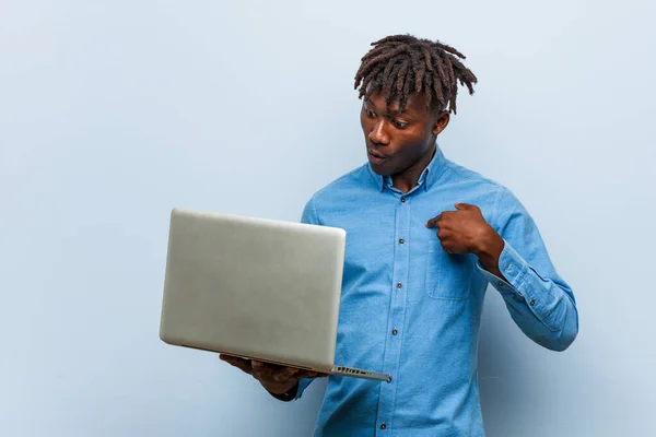Young rasta black man holding a laptop surprised pointing at himself, smiling broadly.