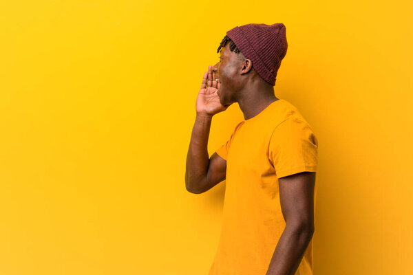 Young black man wearing rastas over yellow background shouting and holding palm near opened mouth.