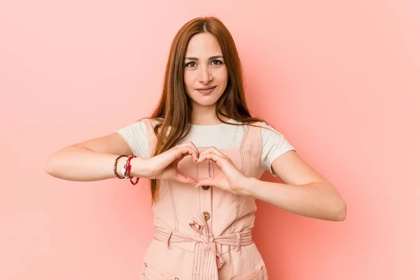 Young ginger woman with freckles smiling and showing a heart shape with hands.