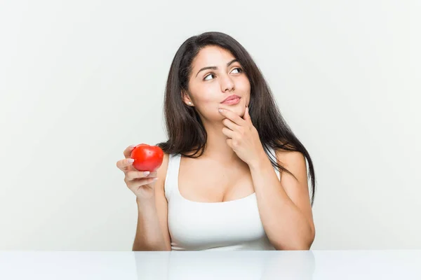 Jovem Hispânica Segurando Tomate Olhando Para Lado Com Expressão Duvidosa — Fotografia de Stock