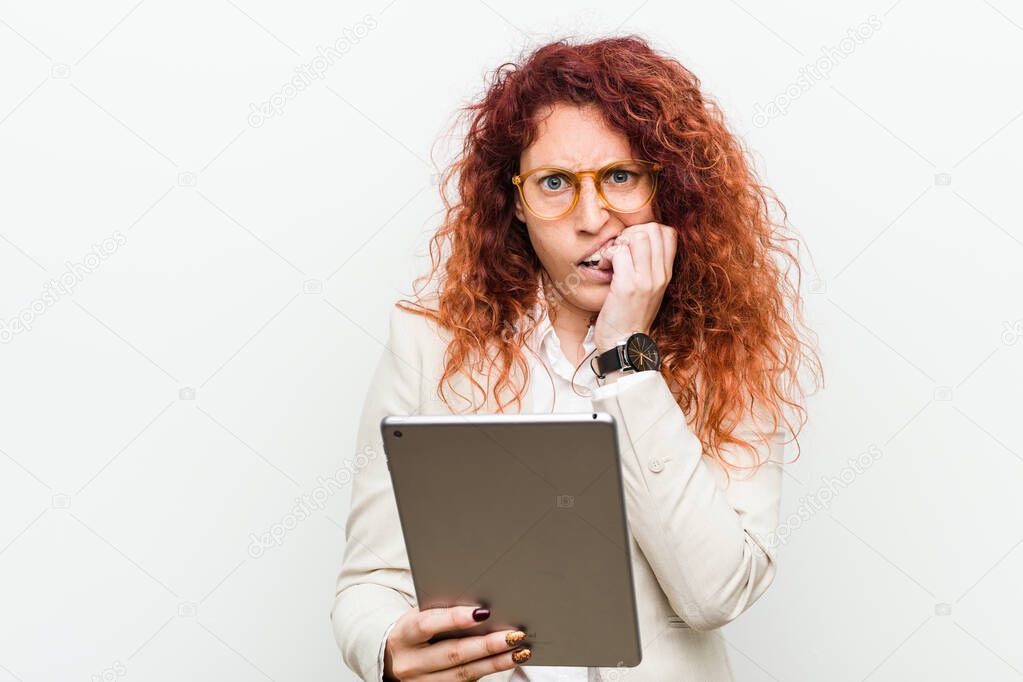 Young caucasian business redhead woman holding a tablet biting fingernails, nervous and very anxious.