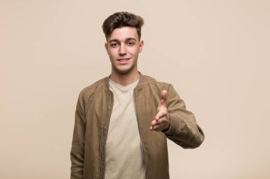 Kahverengi ceketli genç beyaz adam selamlama jestinde kameraya elini uzatıyor..