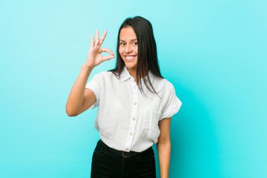 Mavi bir duvara yaslanmış genç, Latin asıllı havalı bir kadın. Neşeli ve kendinden emin..