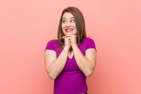Young hispanic woman against a pink wall keeps hands under chin, is looking happily aside.