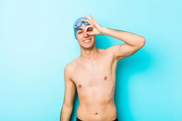 Young swimmer man excited keeping ok gesture on eye.