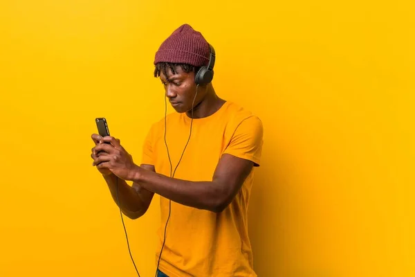 Young african man standing against a yellow background wearing a hat listening to music with a phone