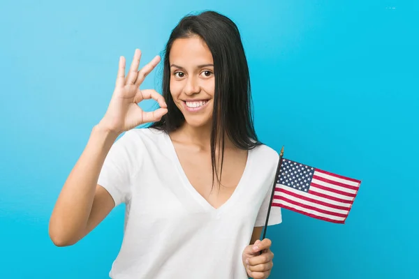 Young hispanic woman holding a united states flag cheerful and confident showing ok gesture.