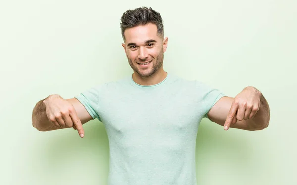 Mladý Pohledný Muž Zeleném Pozadí Ukazuje Prsty Dolů Pozitivní Pocit — Stock fotografie