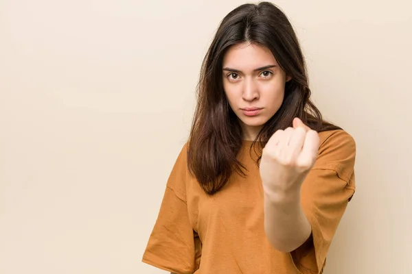 Young brunette woman against a beige background showing fist to camera, aggressive facial expression.