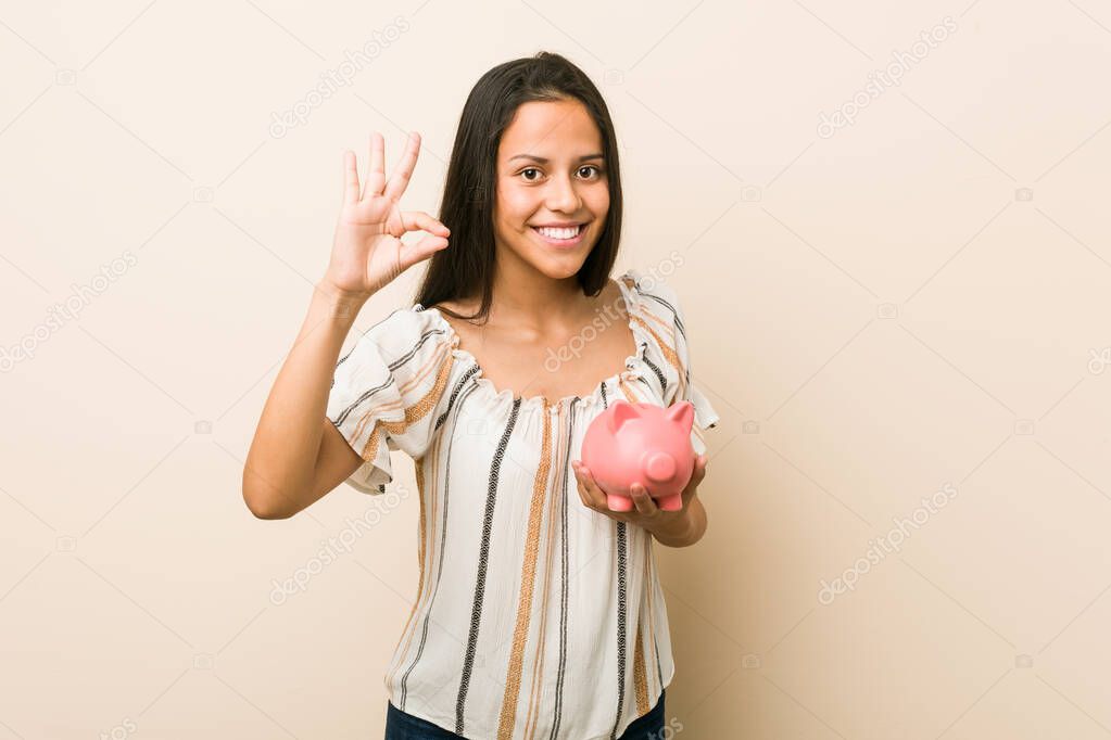 Young hispanic woman holding a piggy bank cheerful and confident showing ok gesture.