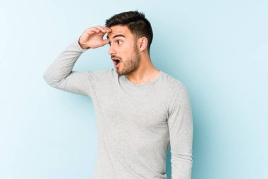 Kafkasyalı genç adam mavi arka planda izole edilmiş, uzaklara bakıyor ve elini alnına koyuyor..