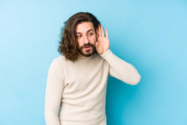 Uzun saçlı genç adam mavi arka planda izole edilmiş bir şekilde bir dedikoduyu dinlemeye çalışıyor..