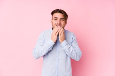 Pembe arka planda poz veren genç beyaz bir adam bir şeylere gülüp, elleri ile ağzını kapatıyor..