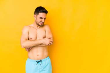 Sarı arka planda mayosu olan genç, beyaz bir adam. Dirseğine masaj yapan beyaz bir genç. Kötü bir hareketin ardından acı çekiyor..