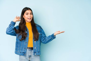 Mavi arka planda izole edilmiş genç bir Hintli kadın avuç içinde fotokopi alanı tutuyor, el üstünde tutuyor. Şaşırmış ve mutlu..