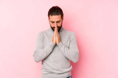 Genç, uzun saçlı, pembe arka planda izole bir adam. El ele tutuşup, ağzının kenarında dua ediyor. Kendine güveniyor..
