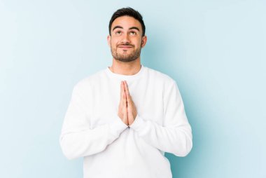 Mavi arka planda izole edilmiş genç beyaz bir adam el ele tutuşup, ağzının kenarında dua ediyor, kendinden emin..