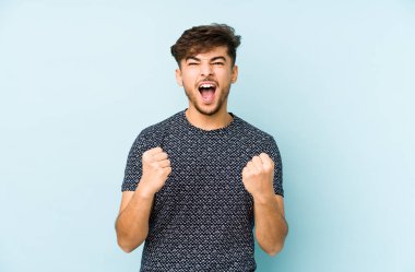 Genç Arap adam mavi bir arka planda izole edilmiş tasasız ve heyecanlı bir şekilde tezahürat yapıyor. Zafer konsepti.