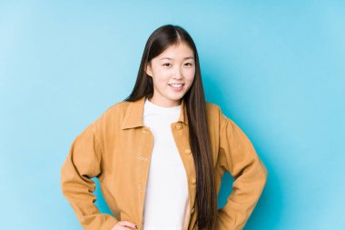 Mavi arka planda poz veren Çinli genç bir kadın izole edilmiş, kendine güveni tam, ellerini kalçasında tutuyor..