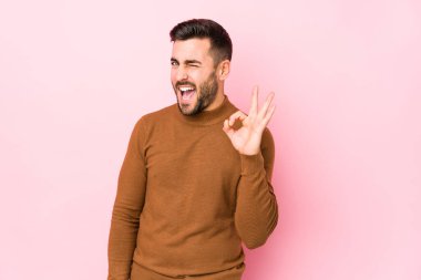 Pembe arka planda genç beyaz bir adam bir göz kırpıyor ve elleriyle iyi bir jest yapıyor..