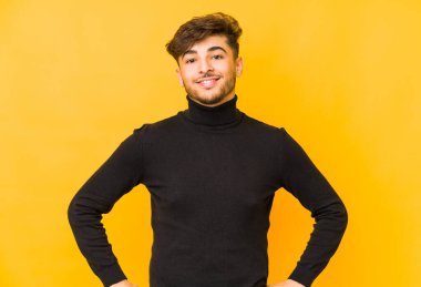 Sarı arka planda izole edilmiş genç bir Arap adam ellerini kalçalarına koyuyor..