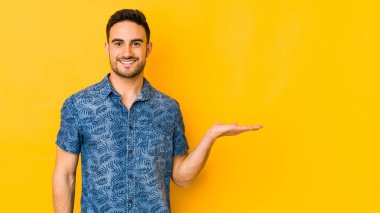 Sarı fırında izole edilmiş genç beyaz bir adam bir palmiye üzerinde fotokopi alanı gösteriyor ve belinde başka bir el tutuyor..