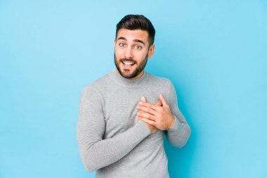 Mavi arka planı olan beyaz bir genç, dost canlısı bir ifadeye sahip, avuçlarını göğsüne bastırıyor. Aşk konsepti.