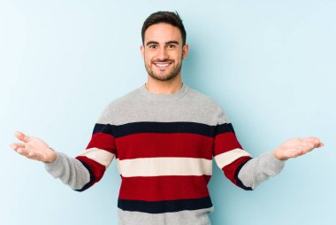 Mavi arka planda izole genç beyaz adam kollarıyla ölçek yapar, mutlu ve kendinden emin hisseder..