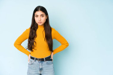 Mavi arka planda izole edilmiş genç Hintli kadın yanakları patlatıyor, yorgun bir ifadesi var. Yüz ifadesi kavramı.