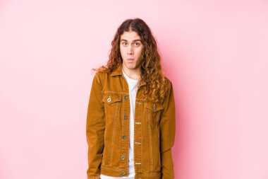 Genç, uzun saçlı bir adam omuz silkiyor, omuzları açık ve gözleri karışık..