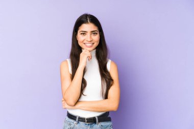 Mor arka planda izole edilmiş genç Hintli kadın mutlu ve kendinden emin gülümsüyor, elleriyle çenesine dokunuyor..