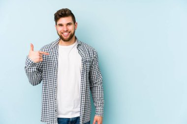 Mavi arka planda izole edilmiş genç beyaz adam bir gömlek fotokopi alanını işaret ediyor, gururlu ve kendinden emin.