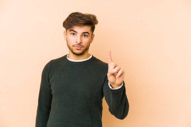 Genç Arap bir adam bej bir arka planda izole edilmiş. Parmağıyla bir numarayı gösteriyor..