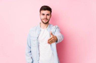 Beyaz arka planda poz veren beyaz tenli genç bir iş adamı kameraya doğru elini uzatarak selam veriyor..