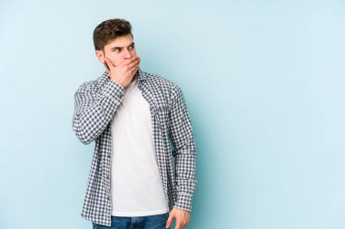 Kafkasyalı genç adam mavi arka planda izole edilmiş. Düşünceli bir şekilde ağzıyla ağzını kapatan bir fotokopi odasına bakıyor..