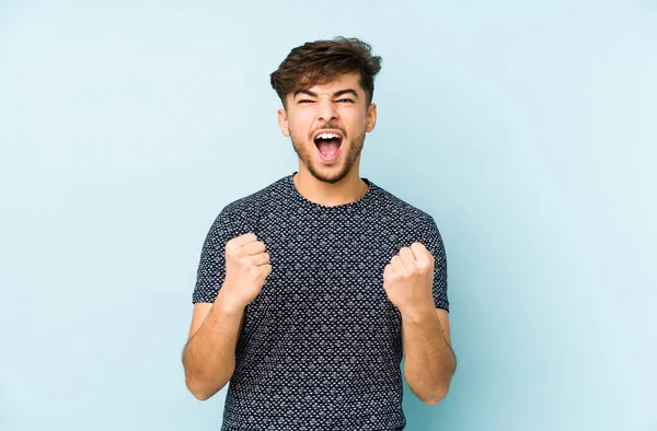 Genç Arap adam mavi bir arka planda izole edilmiş tasasız ve heyecanlı bir şekilde tezahürat yapıyor. Zafer konsepti.