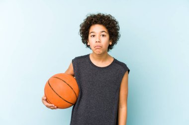 Mavi arka planda izole edilmiş basketbol oynayan çocuk omuzlarını silker ve gözleri açık kalır..