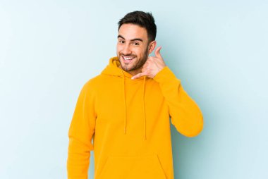 Mavi arka planda izole edilmiş genç beyaz adam parmaklarıyla bir cep telefonu hareketi gösteriyor..