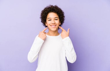 Afro-amerikalı küçük çocuk yalnız gülümsüyor, parmaklarını ağzına doğrultuyor..