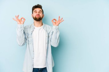 Mavi arka planda izole edilmiş genç beyaz adam yorucu bir günün ardından rahatlıyor. Yoga yapıyor..