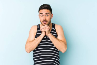 Kafkasyalı genç adam mavi arka planda izole edilmiş şans için dua ediyor, şaşırmış ve ağzını açıp önüne bakıyor..