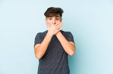 Mavi arka planda izole edilmiş genç bir Arap adam elleriyle ağzını kapatırken şok oldu..