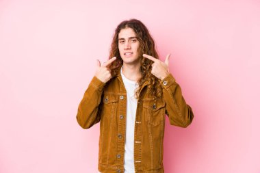 Uzun saçlı genç adam, yalnız gülümsüyor, parmaklarını ağzına doğrultuyor..