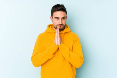 Kafkasyalı genç adam mavi arka planda izole edilmiş dua ediyor, bağlılık gösteriyor, dindar biri ilahi ilham arıyor..