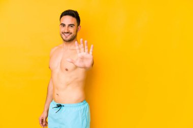 Sarı arka planda mayosu olan genç beyaz bir adam. Beş numarayı parmaklarıyla gülümseyen beyaz bir adam..