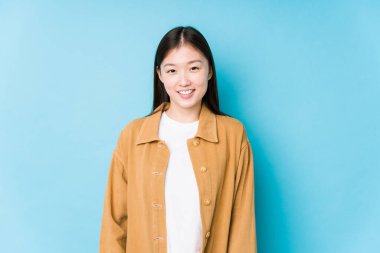 Mavi arka planda poz veren genç Çinli kadın mutlu, güler yüzlü ve neşeli..