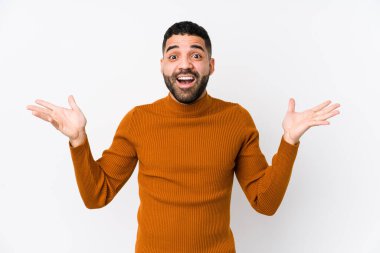 Genç Latin bir adam beyaz bir arkaplanda zaferi ya da başarıyı kutluyor, şaşırıyor ve şok oluyor..