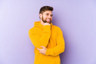 Mor arka planda izole edilmiş genç adam gülümsüyor mutlu ve kendinden emin, elleriyle çeneye dokunuyor..