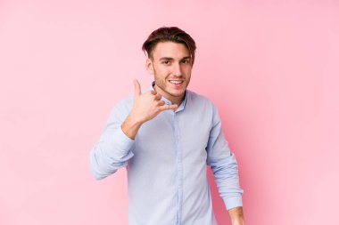 Pembe arka planda poz veren genç beyaz bir adam izole edilmiş, parmaklarıyla bir cep telefonu çağrısı hareketi gösteriyor..