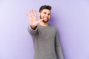 Mor arka planda izole edilmiş genç beyaz adam gülümseyerek beş numarayı parmaklarıyla gösteriyor..